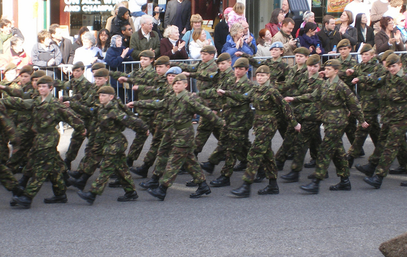 A group of soldiers is shown marching down the road while spectators look on.