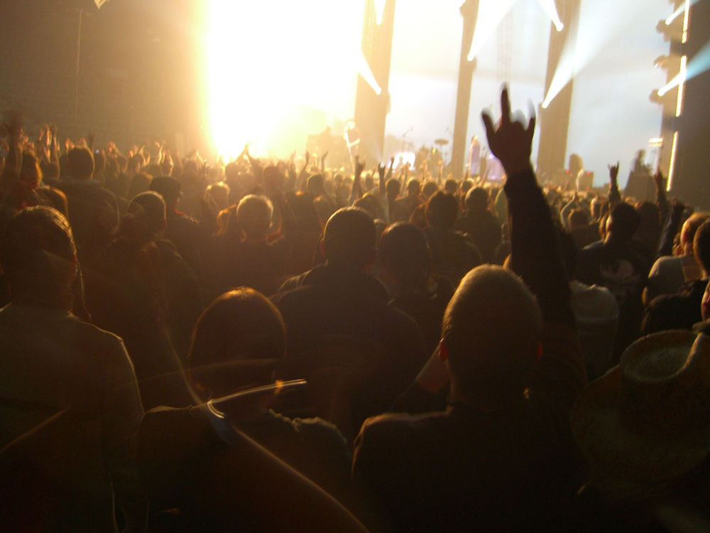A crowd of people at a concert is shown from behind. Multiple lighting effects can be seen emanating from the stage.