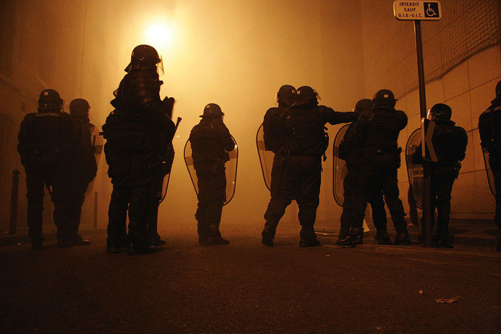 A group of police holding shields are shown standing together at night.