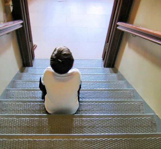 A child is shown from behind sitting on metal stairs looking into a room. 