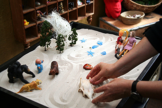 This photograph shows a person playing with objects in a small box filled with sand. The person is organizing these objects and small play figures in a form of treatment called sandplay.