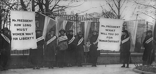 A photograph shows Alice Paul and the Silent Sentinels picketing outside of the White House. Each woman wears a banner stating her alma mater. The women hold two large signs, the first of which reads 