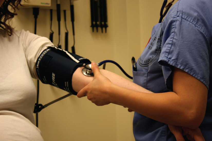 This photo shows a nurse taking a woman’s blood pressure with a blood pressure cuff. The nurse is pumping the cuff with her right hand and holding a stethoscope on the patient’s arm with her left hand.