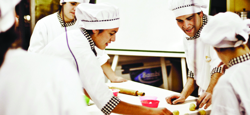 A photo of several bakers at work on a table in a classroom setting.