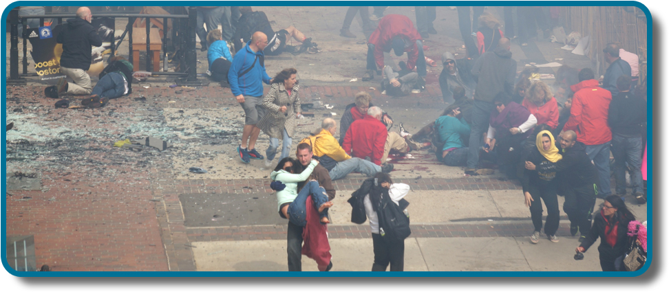 A photograph shows a crowd  at the site of the Boston Marathon bombing immediately after it occurred. Debris is scattered on the ground, several people appear to be injured, and several people are helping others.