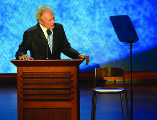 An image of Clint Eastwood standing behind a podium. Next to him on the right is an empty chair.