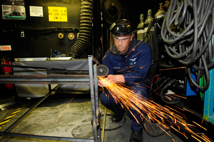 Figure is a photo of a man sharpening a piece of metal on the rotating grindstone. Sparks from the grindstone are clearly evident.