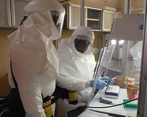 A photograph of two individuals in white biohazard suits that completely covers the individual, complete with head and face covering. They are working in a hood.